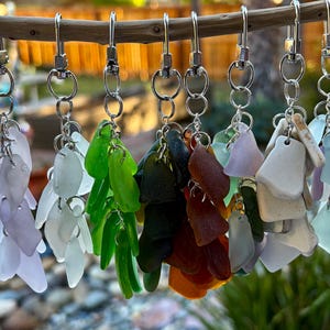May include: A collection of nine keychains featuring clusters of multi-colored sea glass pieces. Each keychain is attached to a silver-toned metal swivel clasp and jump rings, showcasing various shades of lavender, green, brown, white, and blue.