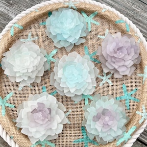 May include: A round decorative tray with a burlap surface, a white rope border, and painted turquoise starfish. The tray holds six layered, translucent, flower-shaped objects in shades of blue, purple, and white.