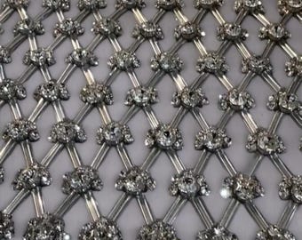 Op de afbeelding: Zilveren mesh-stof met een herhalend patroon van heldere strass steentjes. De strass steentjes zijn gerangschikt in een diamantvorm en zijn gezet in een zilveren metalen frame.