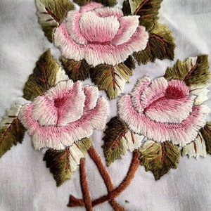 Può includere: Primo piano di rose ricamate con petali rosa e bianchi e foglie verdi su uno sfondo di tessuto bianco.