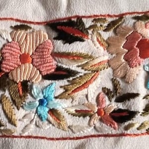 May include: A white fabric with a decorative embroidered border. The border features a floral design with pink, orange, and green flowers. The flowers are embroidered with a variety of stitches, including satin stitch and French knots.