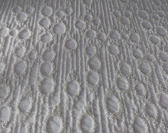 Op de afbeelding: Witte stof met een patroon van verticale lijnen en cirkelvormige ontwerpen, allemaal versierd met kleine, glanzende kralen. De kralen creëren een gestructureerd, elegant oppervlak, geschikt voor formele kleding of decoratieve projecten.