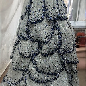 May include: A strapless white gown with a layered skirt, embellished with silver and navy blue sequins. The bodice is adorned with silver sequins, and a white sash accentuates the waist. The dress is displayed on a mannequin.