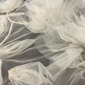 Puede incluir: Primer plano de una delicada tela de tul de color blanco roto con un dise&ntilde;o floral. La tela est&aacute; en capas y fruncida, creando un aspecto suave y rom&aacute;ntico.