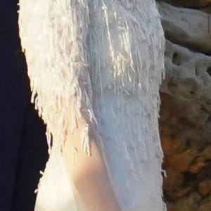 May include: A white wedding dress with a unique fringed design on the sleeves and bodice. The dress appears to be made of a lightweight fabric, creating a flowing silhouette. The fringing adds texture and movement.