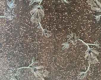 Puede incluir: Un primer plano de una tela marrón claro con un delicado patrón floral. La tela está cubierta de pequeñas lentejuelas iridiscentes que crean un efecto brillante. El diseño floral presenta flores blancas con un sutil tono azul, y los tallos están adornados con pequeñas cuentas brillantes.