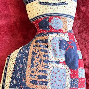 May include: A handmade cat-shaped pillow with a patchwork design. The pillow features a combination of blue, red, and cream fabrics with floral and polka dot patterns. The cat's face has button eyes and a stitched mouth. The pillow is a decorative item.