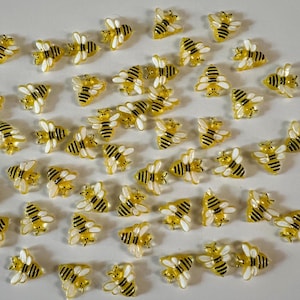 May include: A collection of small, yellow and black bee-shaped embellishments. Each bee has white wings and is arranged randomly. These decorative accents are suitable for crafts and DIY projects.
