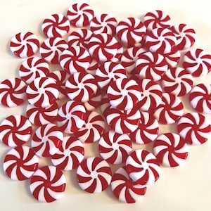 May include: A collection of small, round, flat peppermint-style candies featuring a red and white spiral pattern. Each piece has a smooth, glossy finish and a circular shape, arranged in a scattered pile on a plain, light-colored surface.