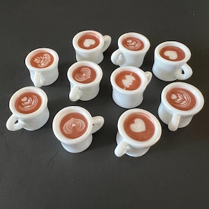 May include: A collection of small, white ceramic coffee cups, each filled with a brown liquid and topped with latte art designs. The latte art includes hearts, swirls, and other patterns. The cups are arranged on a dark surface.