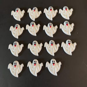 May include: A collection of sixteen white ghost-shaped embellishments with black eyes and red tongues. The ghosts are arranged in four rows of four on a black background. These are likely for Halloween crafts.