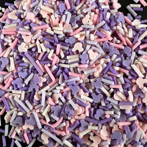 May include: A close-up view of colorful sprinkles in various shapes and sizes. The sprinkles are in shades of pink, purple, and white. Some sprinkles are shaped like Mickey Mouse, while others are small rods. The sprinkles are scattered.