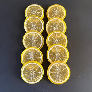May include: Ten yellow lemon slice coasters with white details are arranged in two columns on a dark surface. The coasters have a realistic design, perfect for protecting tables from drink condensation.