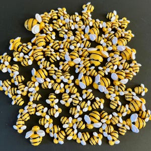 May include: A collection of small, yellow and black bee-shaped embellishments with white wings. The bees are scattered across a dark surface, creating a playful and decorative arrangement. These miniature bees are ideal for crafting and adding a whimsical touch.
