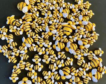 May include: A collection of small, yellow and black bee-shaped embellishments with white wings. The bees are scattered across a dark surface, creating a playful and decorative arrangement. These miniature bees are ideal for crafting and adding a whimsical touch.