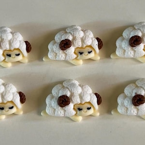 May include: Ten small, white sheep figurines with brown accents and smiling faces are arranged in two rows. The sheep have fluffy wool and are in a cartoon style. The figurines are on a light-colored surface.