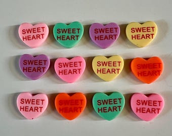 May include: An assortment of colorful heart-shaped candies. Each candy has the words "SWEET HEART" printed on it in a contrasting color. The candies are in shades of pink, green, purple, yellow, and orange.