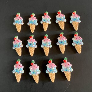 May include: A collection of twelve ice cream cone-shaped embellishments. Each cone has a waffle texture and is topped with pink and blue ice cream, sprinkles, and a red cherry. The embellishments are arranged on a dark surface.