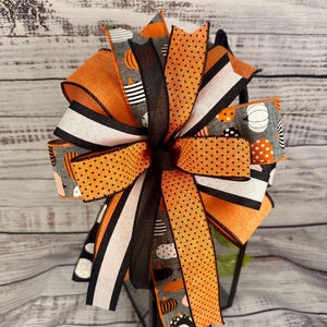 May include: A Halloween-themed bow made with orange, black, and white ribbon. The ribbon features a polka dot pattern, stripes, and a pumpkin print.