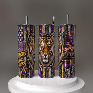 Puede incluir: Tres vasos cilíndricos con pajitas de metal que presentan un patrón de bandera desgastada en morado y dorado. El diseño incluye una cara de tigre, el texto LSU, GEAUX TIGERS y un gráfico de estadio. Los vasos muestran una huella de pata.