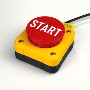 May include: A square industrial push button switch featuring a bright red circular button with the word START printed in white. The base is yellow with black sides and is secured by four silver screws, with a black electrical cable attached.