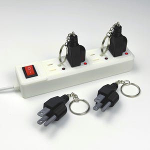 May include: A white electrical power strip with a red power switch and four outlets, two of which contain black electrical plug keychains. Two additional black electrical plug keychains are positioned in front of the power strip on a white surface.