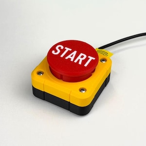 May include: A square industrial push button switch featuring a large, circular red button with the word START printed in white. The housing is yellow with a black base, secured by four silver screws, and includes a black electrical cable attached.