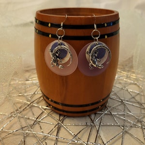 May include: A pair of dangle earrings featuring a silver lizard charm on a purple and pink circular background. The earrings are suspended from silver hooks. The earrings are displayed in front of a wooden barrel.