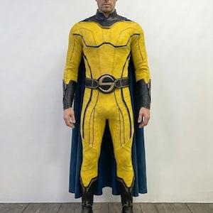 May include: A full-body superhero costume featuring a textured yellow bodysuit with black accents, a high black collar, and a dark blue cape. The ensemble includes black gauntlets, black boots, and a belt with a prominent circular gold S emblem.