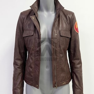 May include: A brown leather jacket featuring a central zipper closure, a high collar, and two chest pockets with flaps. The left sleeve displays a circular orange and red Rebel Alliance emblem. The jacket has long sleeves with zippered cuffs.