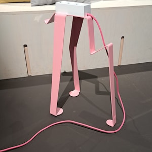 以下が含まれることがあります： ピンクの金属製のスタンドに白い電源タップが置かれています。スタンドは歩く人のようなユニークなデザインです。ピンクのコードが電源タップに接続され、床に置かれています。