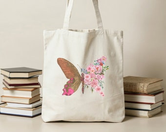 May include: Cream-colored tote bag featuring a butterfly design. One wing is a brown and pink butterfly wing, the other is a collection of pink flowers. The bag is held by a hand, with stacks of books in the background.
