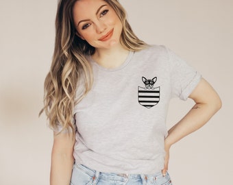 May include: A light gray t-shirt featuring a black and white striped pocket design with a small dog illustration. The dog is wearing glasses. The shirt has rolled-up sleeves and is worn by a person.