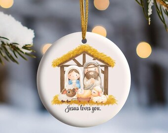 May include: A white ceramic Christmas ornament with a watercolor illustration of the nativity scene. The ornament features Mary, Joseph, and baby Jesus in a manger with two sheep. The text "Jesus loves you." is printed below the scene.