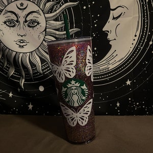May include: A pink glitter tumbler with a white butterfly design and a green Starbucks logo. The tumbler has a clear lid and a green straw.