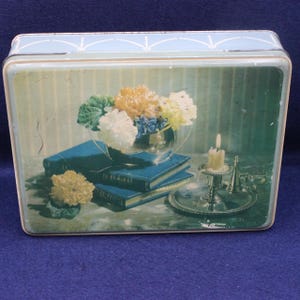 Puede incluir: Fiambrera de metal vintage con una escena de naturaleza muerta floral pintada. La imagen presenta un cuenco plateado con flores, libros azules, una vela encendida y una pequeña flor amarilla. La fiambrera tiene una tapa azul claro con un patrón decorativo.