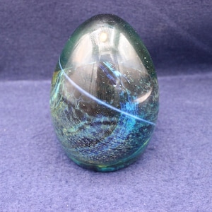 May include: A decorative glass egg with a swirling blue and green design. The egg-shaped object has a glossy finish and a light blue streak across the surface. The egg is set against a dark blue background.