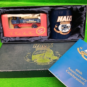 Op de afbeelding: Een geschenkset met een rode en blauwe speelgoedvrachtwagen, een marineblauwe mok met "HALLS"-merk en een boekje. De vrachtwagen is een "Foden Steam Wagon". De set viert een 60-jarig jubileum.