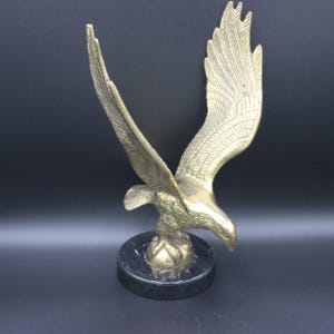 May include: A brass eagle sculpture with outstretched wings, mounted on a round black marble base. The eagle is detailed with textured feathers and a downward gaze, showcasing a classic design. The sculpture is approximately 10 inches tall.