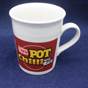 May include: White ceramic mug with a curved handle. The mug features a red and yellow graphic with the words "Golden Wonder Chilli Pot with Rice". The mug is cylindrical and has a smooth, glossy finish.
