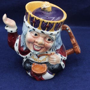 May include: A whimsical ceramic teapot shaped like a king, featuring a smiling face, a purple crown, and a red and white robe. The handle and spout are brown, and the lid is purple with a gold knob. The teapot is a unique decorative item.