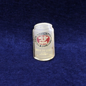 May include: A gold-colored beer can pin with the text "23 Export" in a red circle, surrounded by text in a circular pattern. The pin also features the words "Here de Luxe" and "blonde fine et pure".