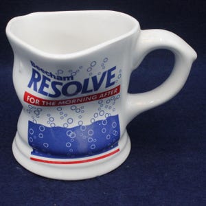 Op de afbeelding: Witte keramische mok met een gebogen handvat en een unieke vorm. De mok heeft de tekst "Beecham RESOLVE FOR THE MORNING AFTER" in blauw en rood, met een blauw en wit bubbelontwerp.