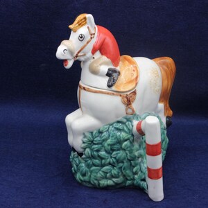 May include: A ceramic horse figurine jumping over a hedge and a striped pole. The horse is white with a brown mane and tail, a red saddle blanket, and a red head covering. The hedge is green, and the pole is white with red stripes.