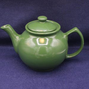 Puede incluir: Una tetera de cerámica verde con tapa y asa a juego. La tetera tiene un cuerpo redondeado, un pico curvo y un asa de bucle. La tapa tiene un pequeño pomo en la parte superior. La tetera es de color verde liso y brillante.