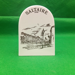 Könnte beinhalten: Weißes, bogenförmiges Keramik-Dekorationsobjekt mit einer Schwarz-Weiß-Illustration einer Straßenszene. Das Wort "Saltaire" und das Jahr "1895" sind oben aufgedruckt. Auf grünem Hintergrund platziert.
