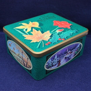 Peut inclure: Boîte en étain rectangulaire verte avec une bordure dorée et un couvercle orné de motifs floraux et saisonniers. Le couvercle présente un narcisse jaune, une feuille d'érable orange et une rose rouge. Les côtés montrent des scènes ovales de paysages d'automne et d'hiver.