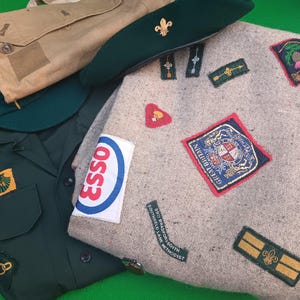 May include: A collection of vintage scout memorabilia. Items include a green beret with a gold emblem, a khaki satchel, and a dark green scout uniform. A gray blanket is adorned with various patches, some with text.