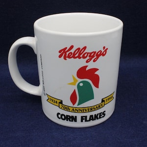 Op de afbeelding: Witte keramische mok met het Kellogg's-logo in rood, een haan-afbeelding met rode, groene en gele accenten en de tekst "70th Anniversary Corn Flakes" met de jaren 1924 en 1994. De mok heeft een gebogen handvat.