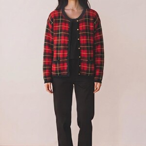 May include: A long-sleeved cardigan featuring a red, black, and yellow plaid pattern with a round neckline, gold-toned buttons, and two small front pockets. The cardigan is worn over a plain black top and black trousers, paired with white sneakers.